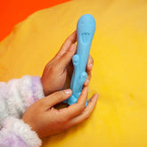 A child's hands are holding a Cute Little Fuckers Trinity - Teal, an irregularly-shaped alien toy from the brand Cute Little Fuckers. The toy, featuring multiple heads and eyes, sits on a yellow surface against an orange background. Its stretchy and playful nature is complemented by whimsical facial features. The child is wearing a fluffy white and purple garment.